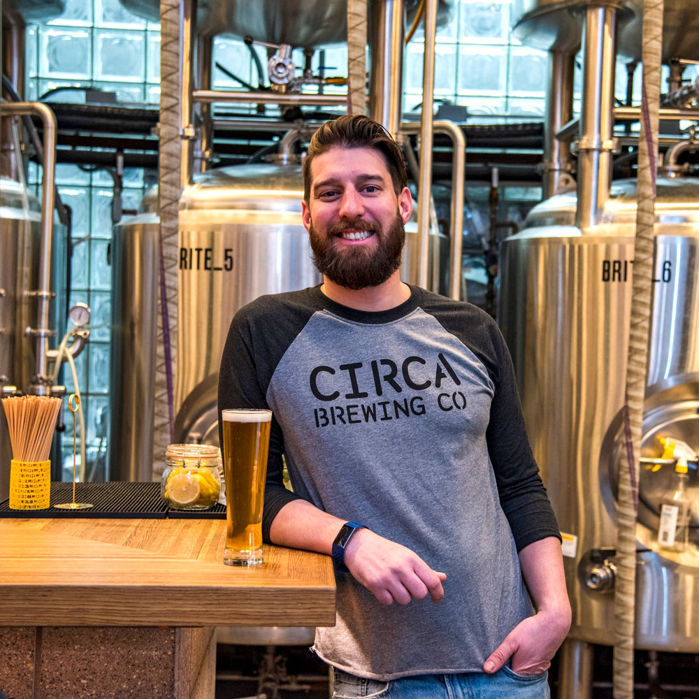 Drew Kostic leaning agaist the bar with a beer glass and breing tanks behind him.